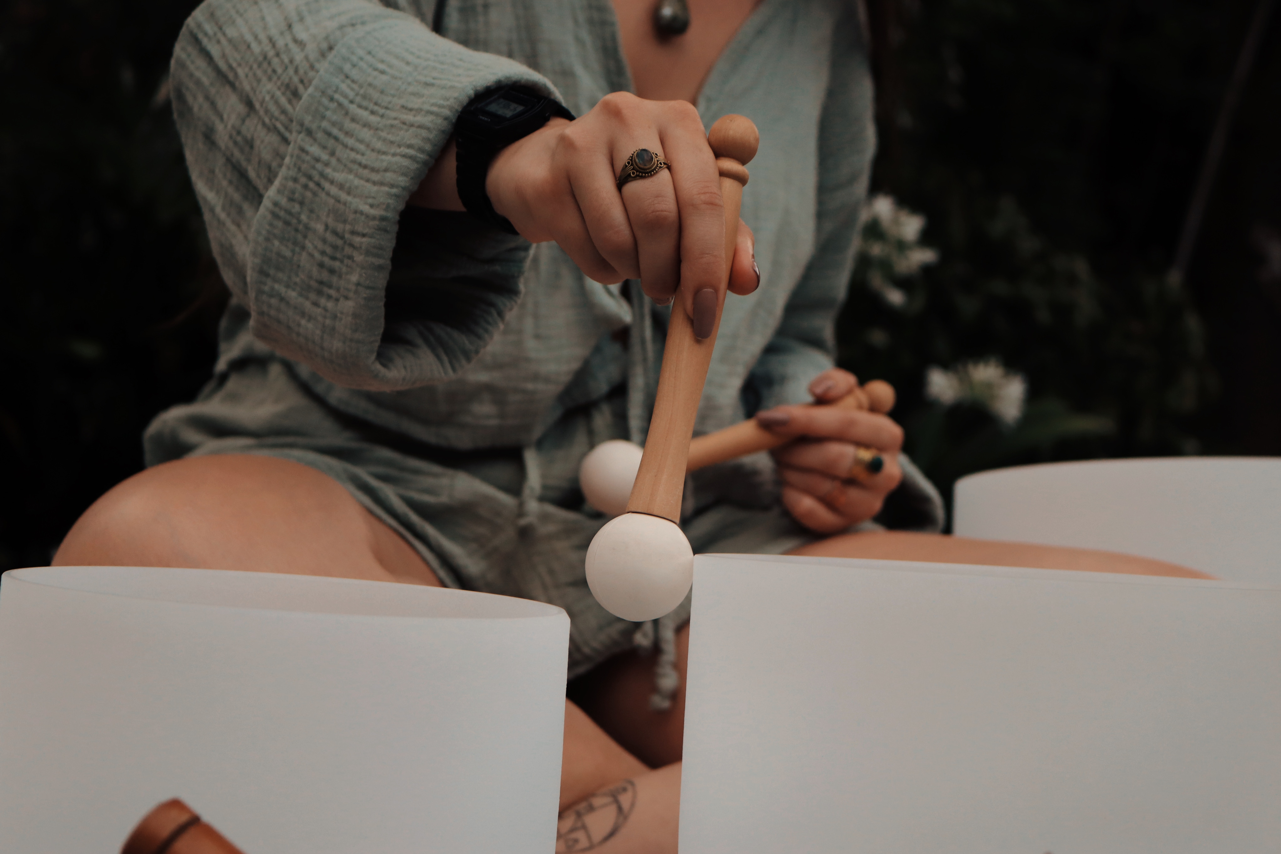 Female wearing green shirt and using her hand to play glass singing bowls with wooden mallets and a garden in the background